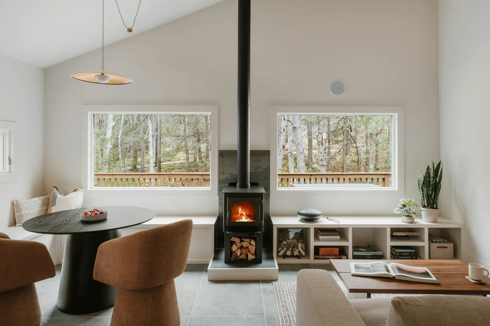 Living room with a black wood stove between two wide windows looking onto trees