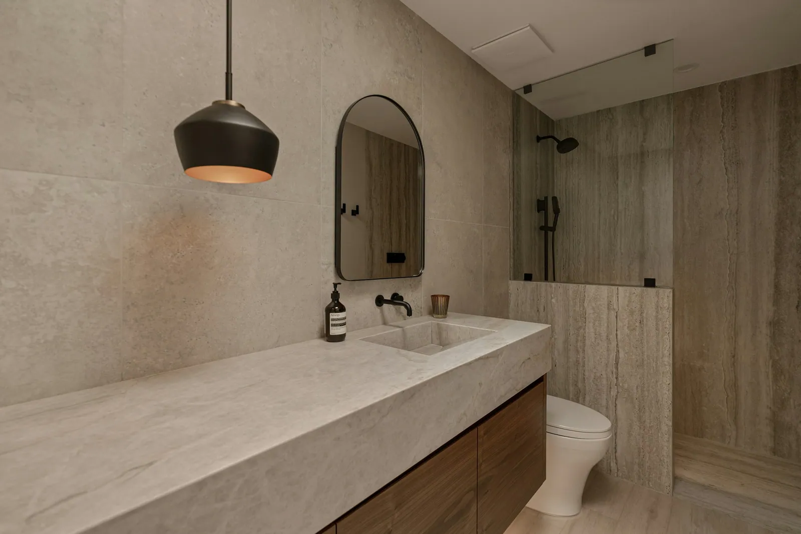 Bathroom with a long stone vanity, wood cabinet and a walk-in shower in travertine