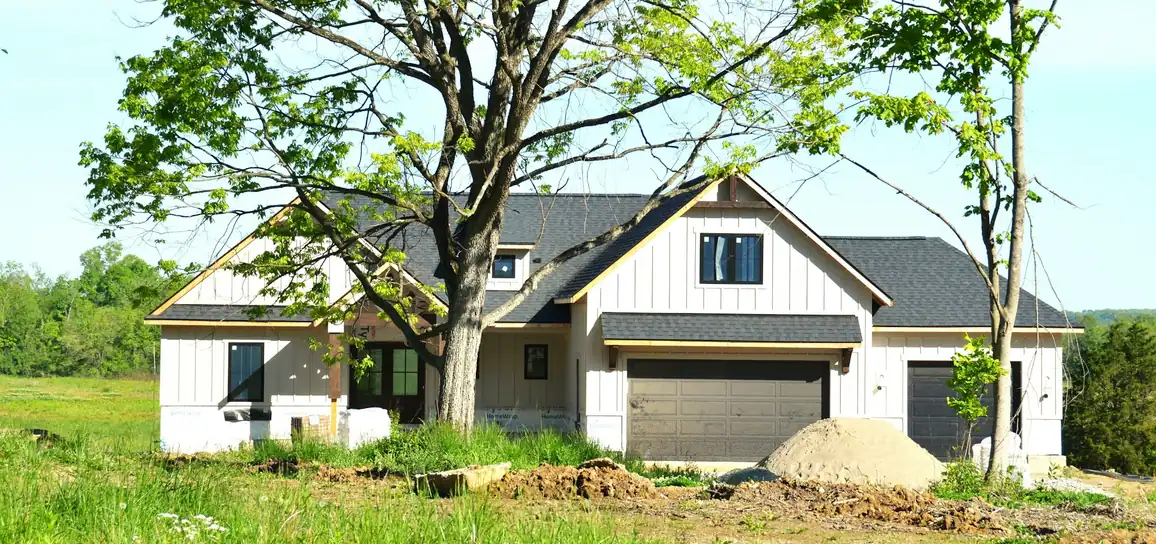 White board-and-batten farmhouse with a dark roof and garage, newly finished, with fresh soil in front